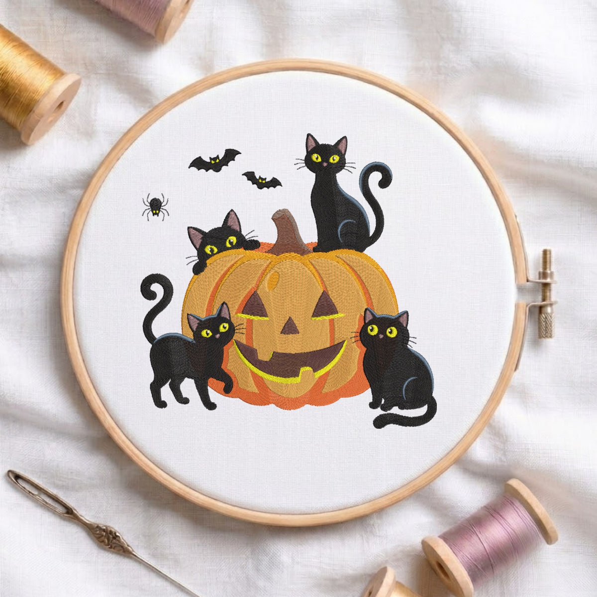 A white fabric stretched in a wooden embroidery hoop, featuring embroidered black cats with glowing yellow eyes surrounding and sitting on top of a carved jack-o'-lantern pumpkin, accented with bats and a spider.