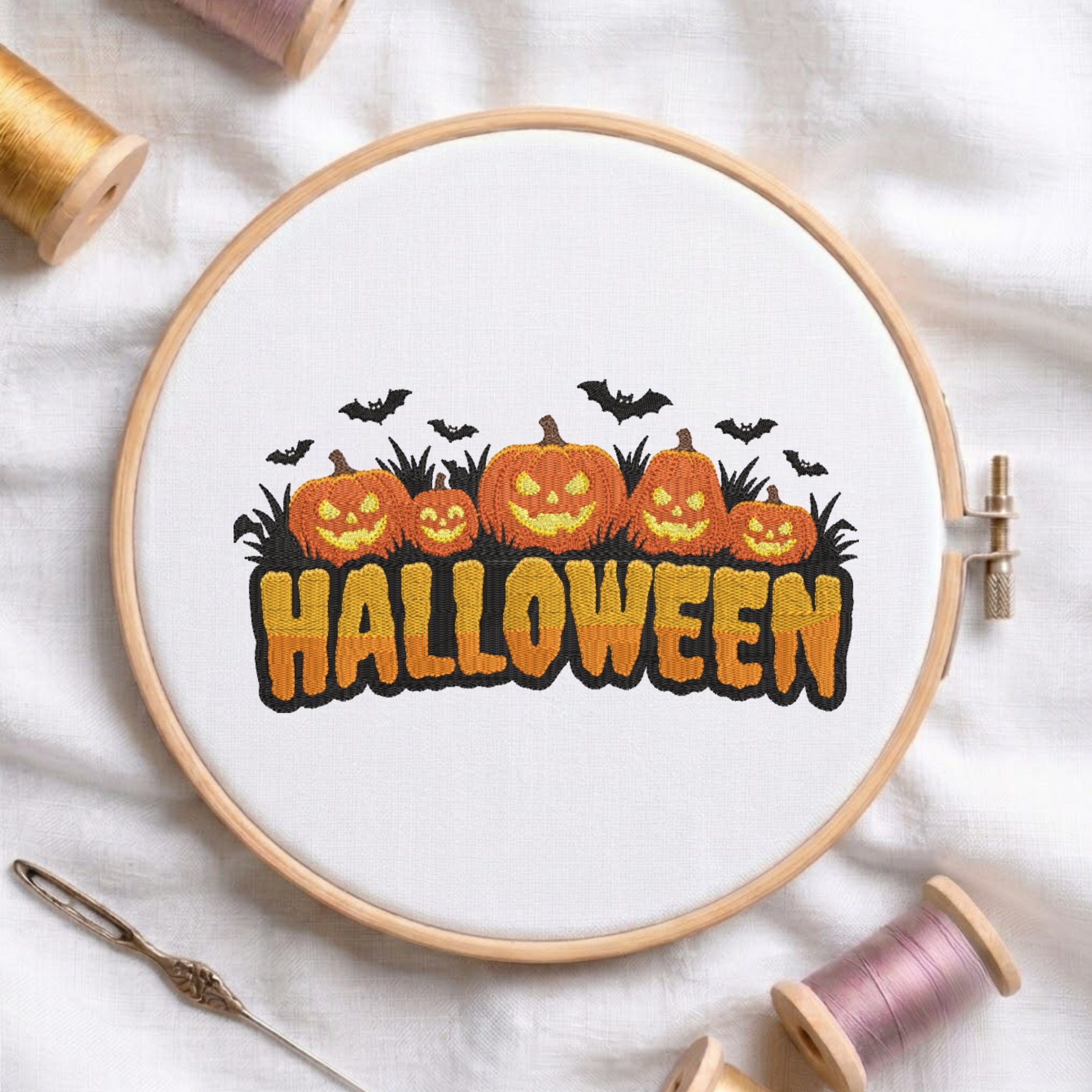 A circular embroidery hoop on white fabric, showing a completed cross-stitch design of orange jack-o'-lantern pumpkins, black bats, and the word 'HALLOWEEN' in orange with black outlines. Spools of thread, a needle, and the embroidery hoop are arranged around it on a white fabric background.