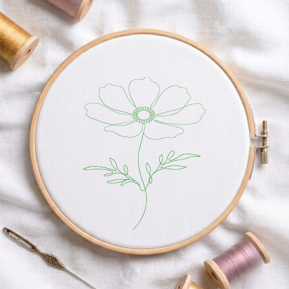 A wooden embroidery hoop holding white fabric with a simple green outline flower design, surrounded by spools of thread and a needle on a white fabric background.