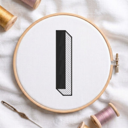 A wooden embroidery hoop holding white fabric with a black outlined letter 'I' design, surrounded by spools of thread and a needle on a white fabric background.