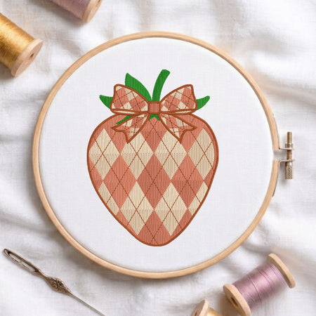 A circular embroidery hoop holding white fabric with a stitched strawberry design featuring an argyle pattern in peach and cream, green leaves, and a matching argyle bow. Spools of thread and a needle are arranged around it on a white fabric background.