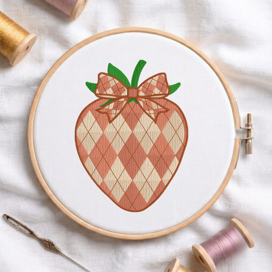A circular embroidery hoop holding white fabric with a stitched strawberry design featuring an argyle pattern in peach and cream, green leaves, and a matching argyle bow. Spools of thread and a needle are arranged around it on a white fabric background.