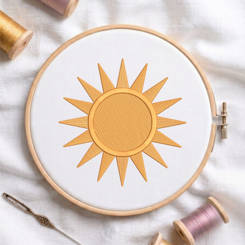 A circular embroidery hoop holding white fabric with a stitched sun motif in warm golden yellow thread. The hoop is styled on a white fabric background with spools of thread and a needle around it.