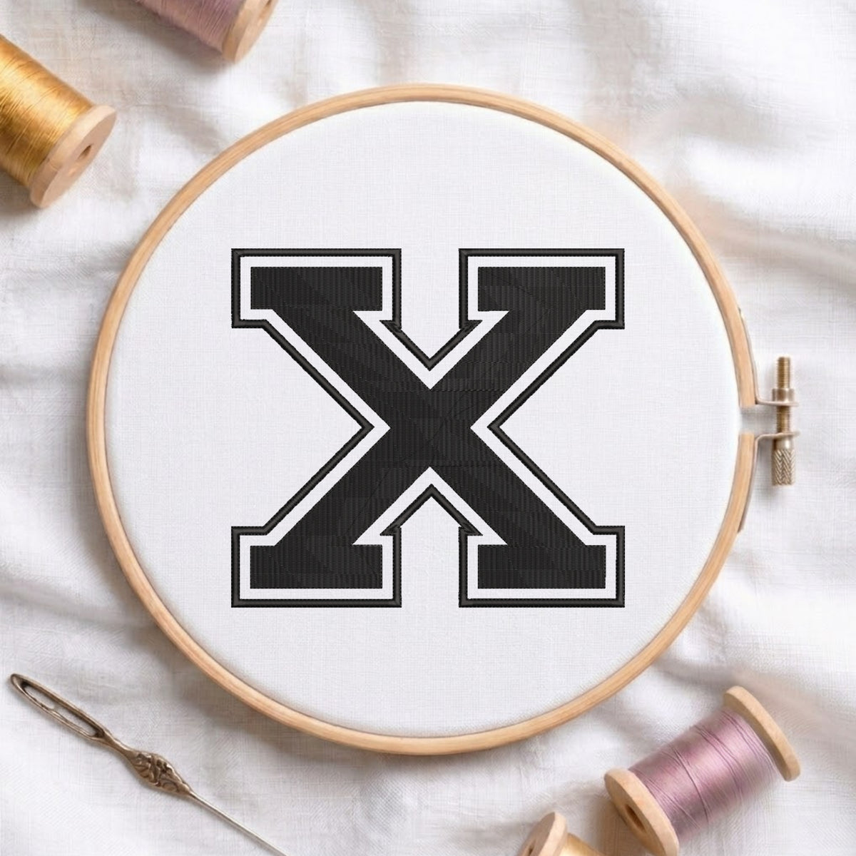 A wooden embroidery hoop holding white fabric with a large black letter X outlined in white. Spools of thread and a needle are arranged around it on a white fabric background.