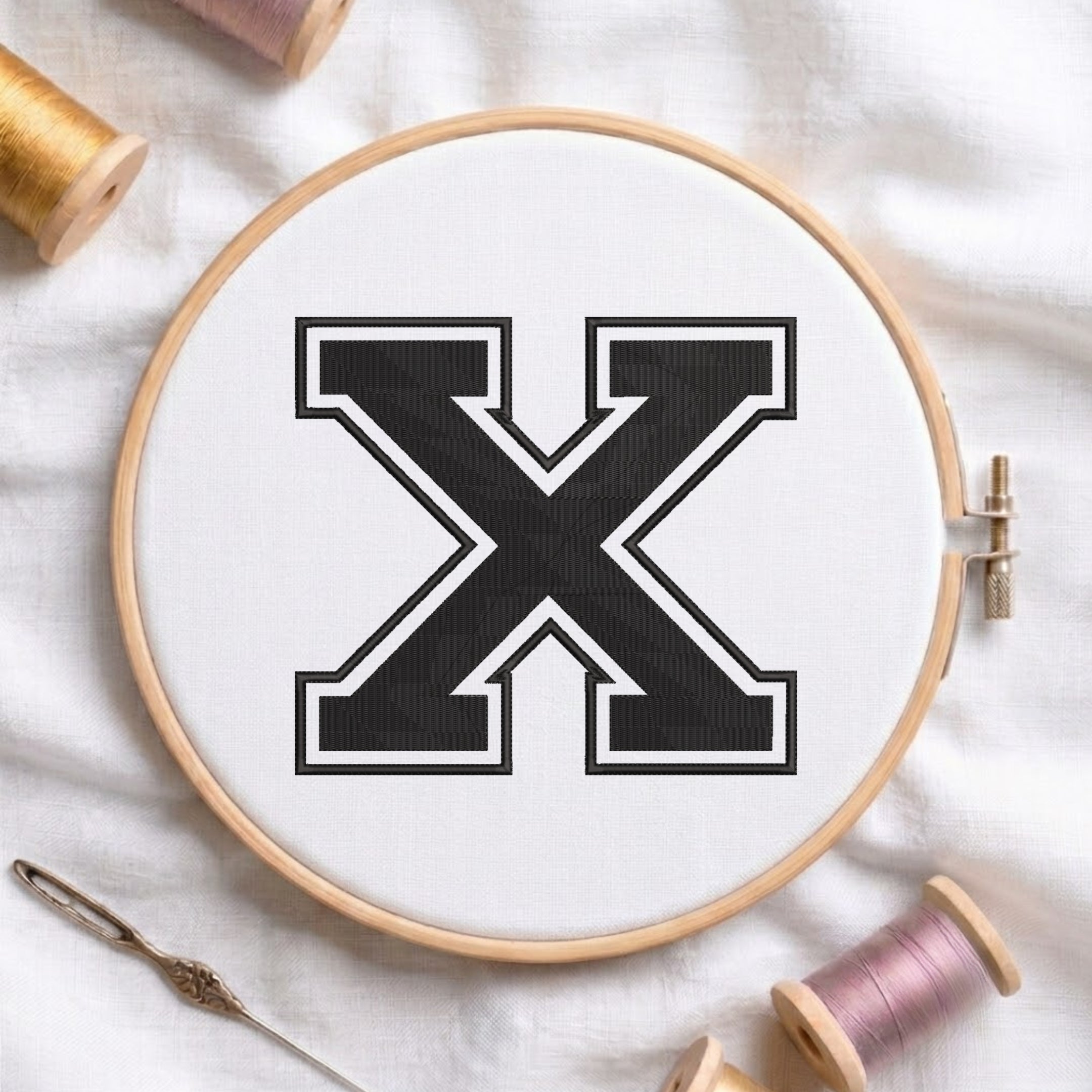A wooden embroidery hoop holding white fabric with a large black letter X outlined in white. Spools of thread and a needle are arranged around it on a white fabric background.