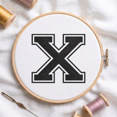 A wooden embroidery hoop holding white fabric with a large black letter X outlined in white. Spools of thread and a needle are arranged around it on a white fabric background.