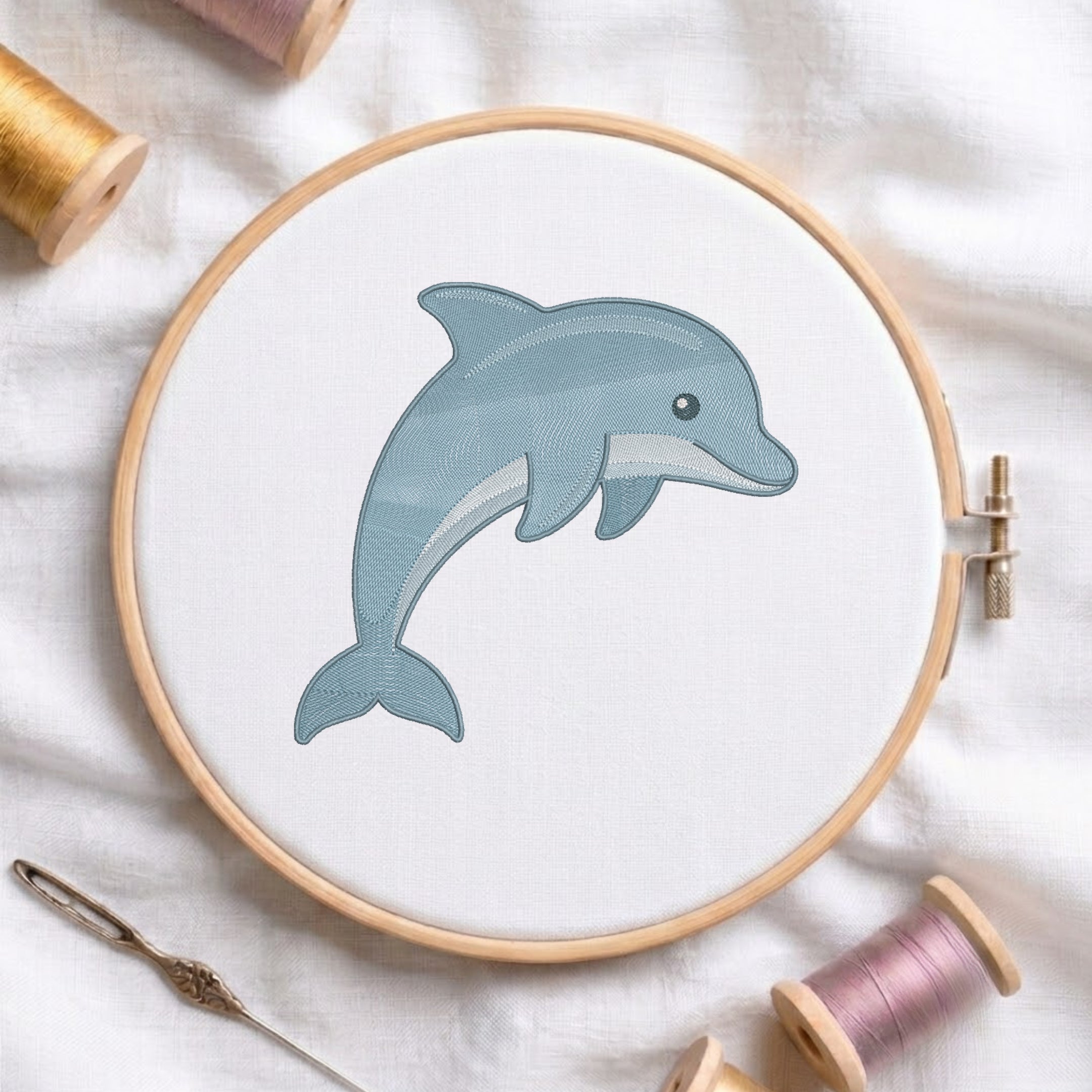 A circular embroidery hoop holding white fabric with a stitched dolphin design in muted blue-gray thread, surrounded by spools of thread and a needle on a white fabric background.