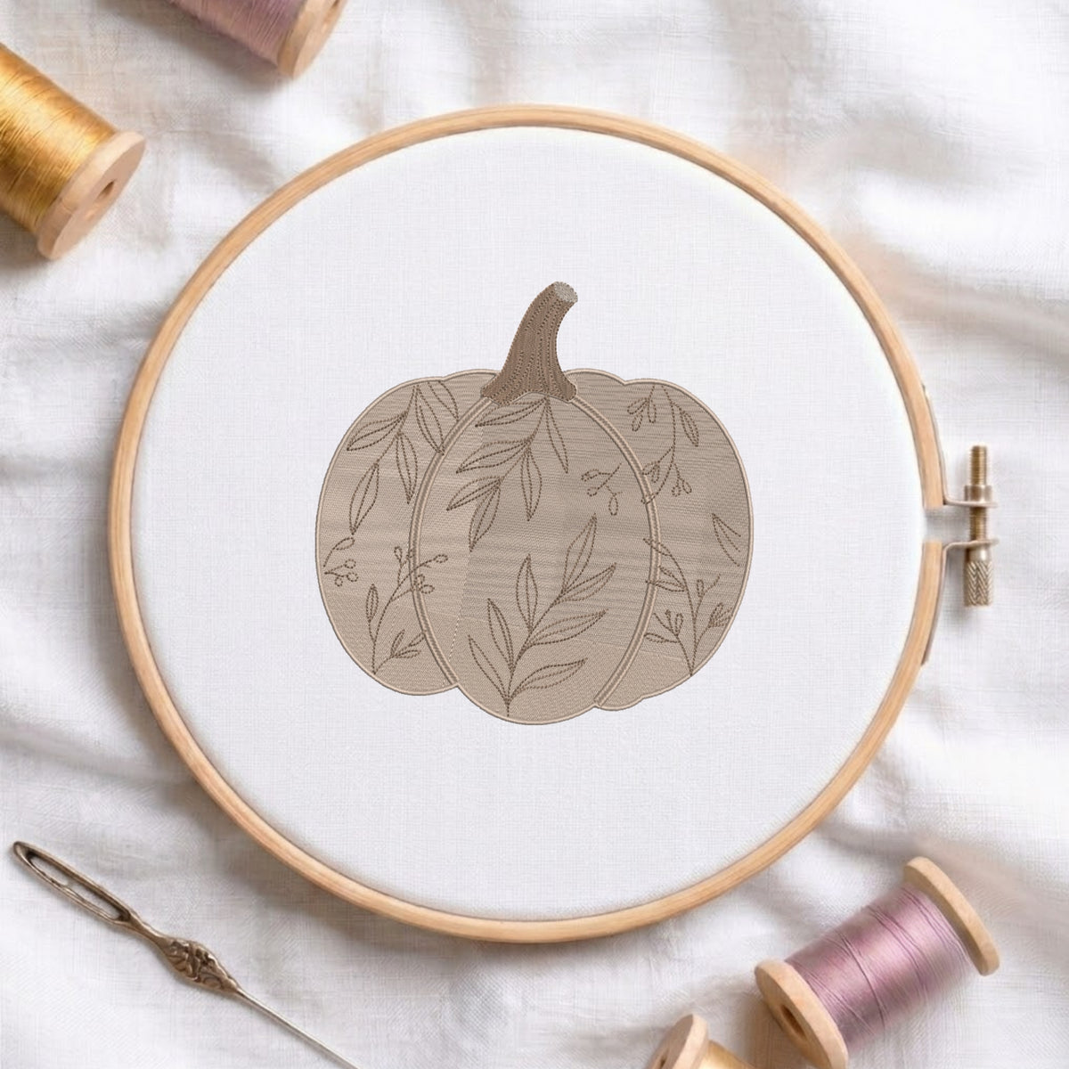 A circular embroidery hoop holding white fabric with a stitched pumpkin design in muted beige and taupe tones. The hoop is styled on a white fabric surface with spools of thread and a needle around it.