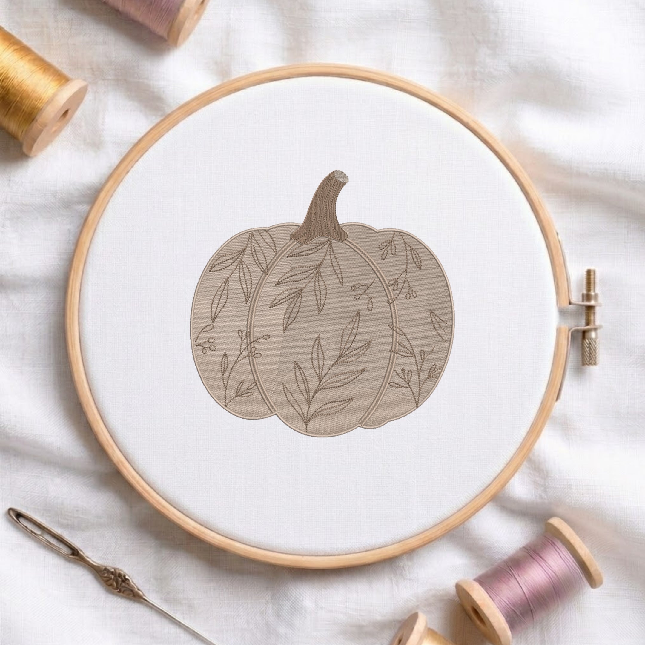 A circular embroidery hoop holding white fabric with a stitched pumpkin design in muted beige and taupe tones. The hoop is styled on a white fabric surface with spools of thread and a needle around it.