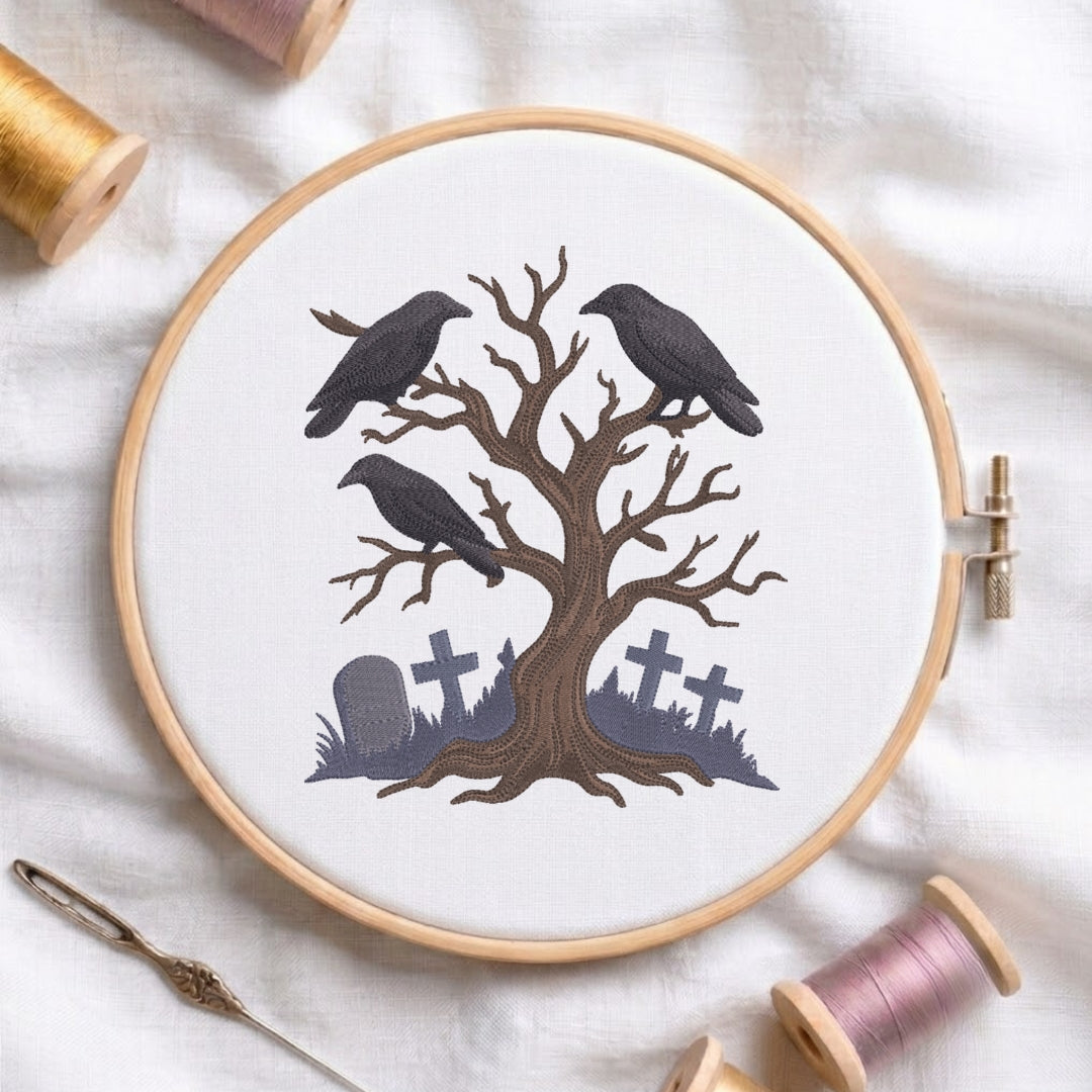 A circular embroidery hoop on white fabric, featuring a stitched design of a bare tree with three black crows and gray tombstones. Spools of thread and a needle are arranged around the hoop.