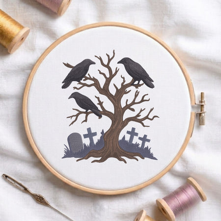 A circular embroidery hoop on white fabric, featuring a stitched design of a bare tree with three black crows and gray tombstones. Spools of thread and a needle are arranged around the hoop.