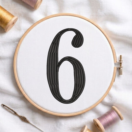 A wooden embroidery hoop holding white fabric with a large black stitched number 6, surrounded by spools of thread and a needle on a white fabric background.