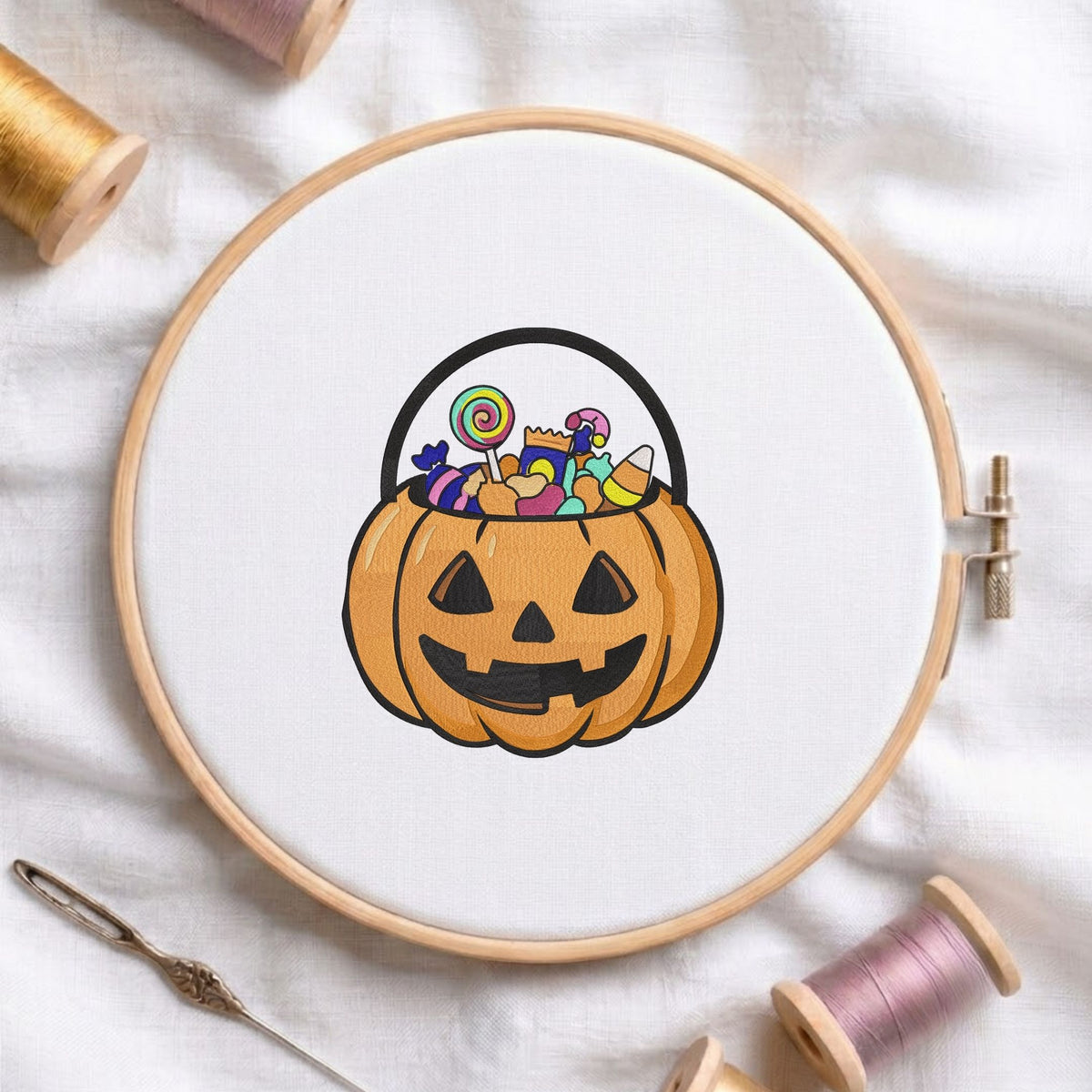 Jack-o'-lantern pumpkin bucket filled with candy and a lollipop machine embroidery design displayed in a wooden embroidery hoop on white fabric with thread spools and a needle