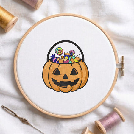 Jack-o'-lantern pumpkin bucket filled with candy and a lollipop machine embroidery design displayed in a wooden embroidery hoop on white fabric with thread spools and a needle