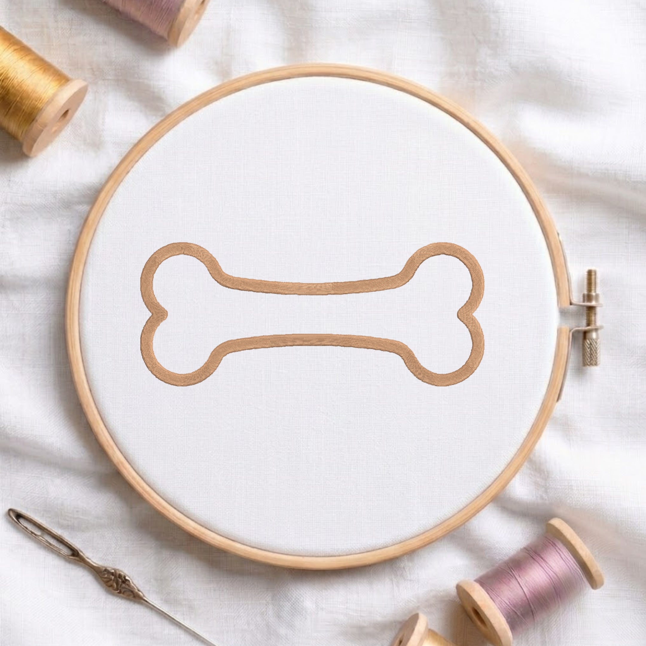 A circular embroidery hoop holding white fabric with a tan bone-shaped embroidery design, styled on a white cloth with spools of thread and a needle around it.