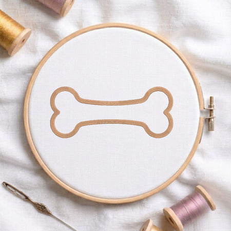 A circular embroidery hoop holding white fabric with a tan bone-shaped embroidery design, styled on a white cloth with spools of thread and a needle around it.