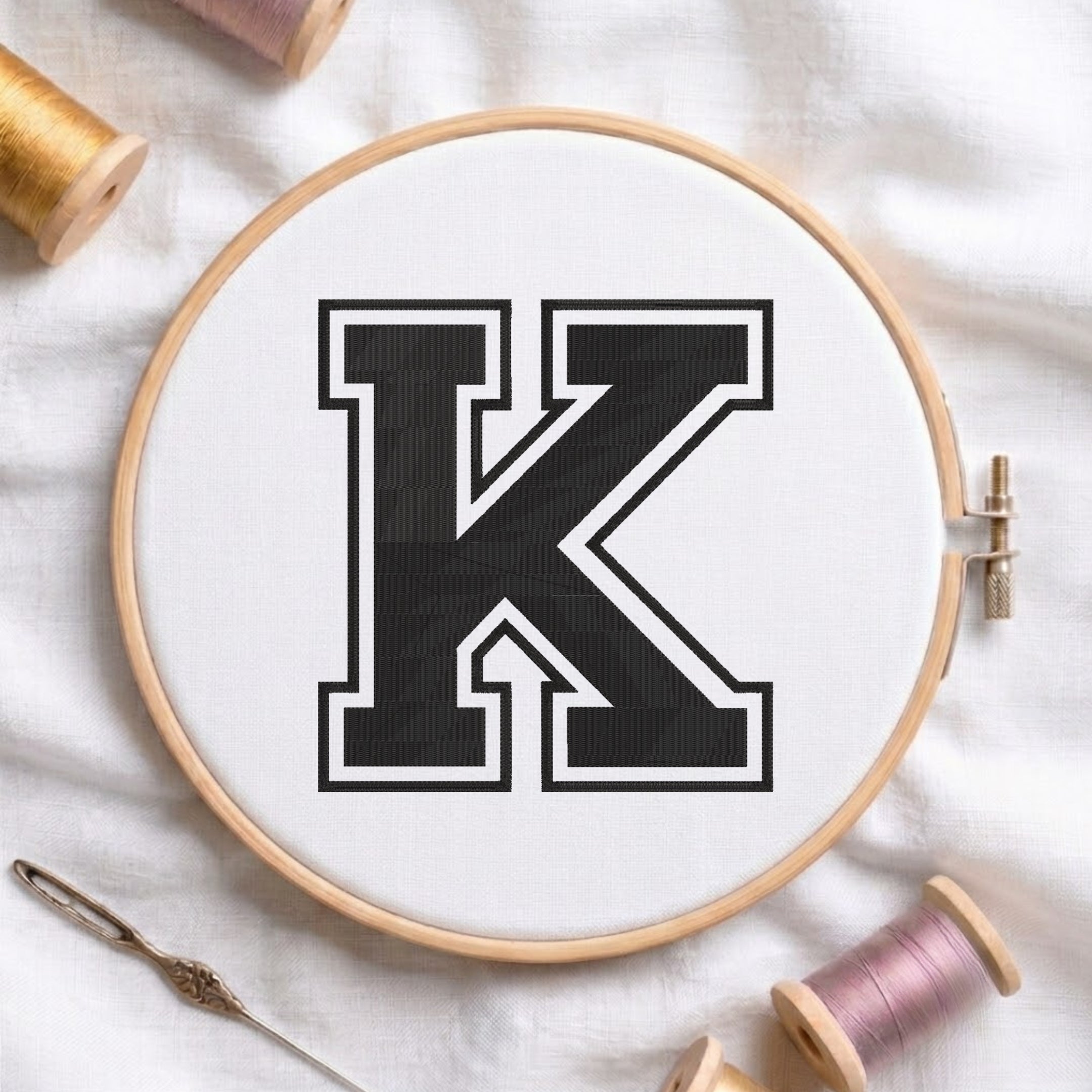 A wooden embroidery hoop holding white fabric with a large black letter K outlined in white, surrounded by spools of thread and a needle on a white fabric background.