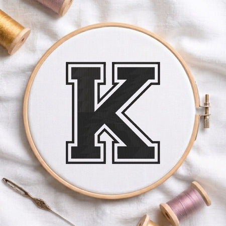 A wooden embroidery hoop holding white fabric with a large black letter K outlined in white, surrounded by spools of thread and a needle on a white fabric background.