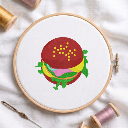 Delicious burger machine embroidery design displayed in a wooden hoop on white fabric with thread spools and a needle