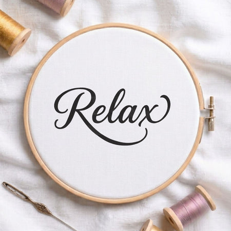 "Relax" script typography machine embroidery design displayed in a wooden hoop on white fabric with thread spools and a needle