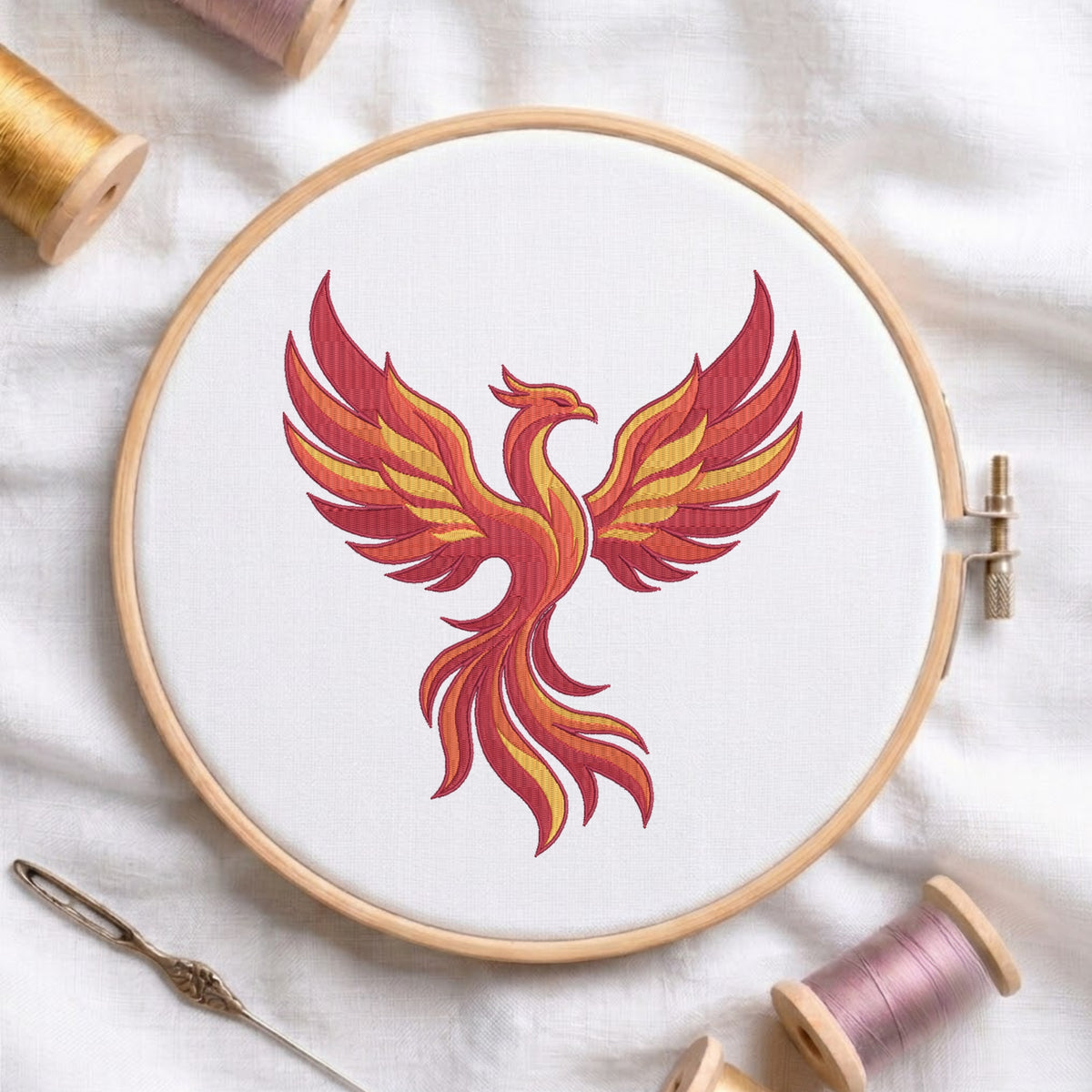 A circular embroidery hoop holding white fabric with a stylized phoenix design in red, orange, and yellow thread. A needle and spools of thread are arranged around it on a white fabric background.