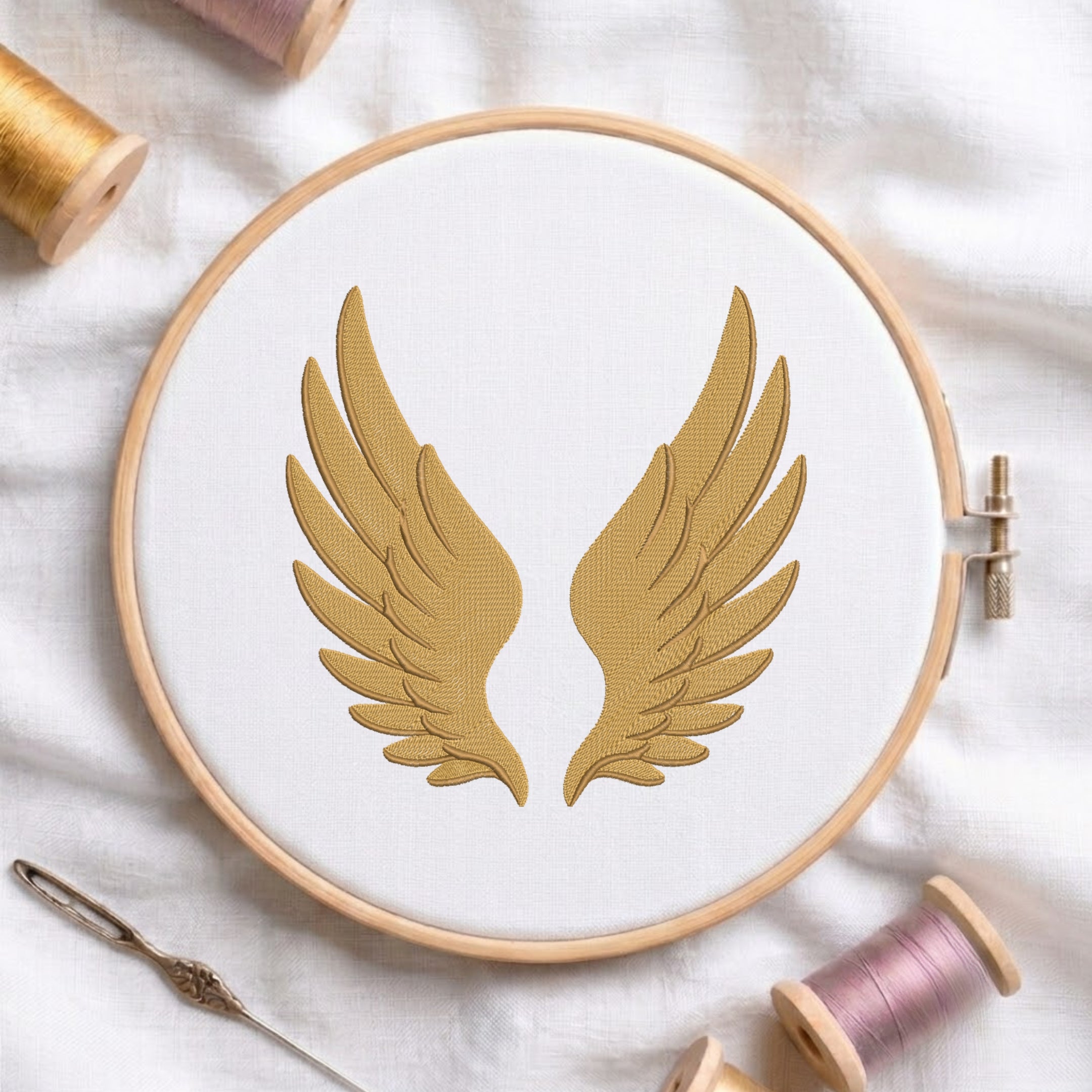 A wooden embroidery hoop holding white fabric with a gold embroidered pair of wings, styled on a white cloth with thread spools and a needle around it.
