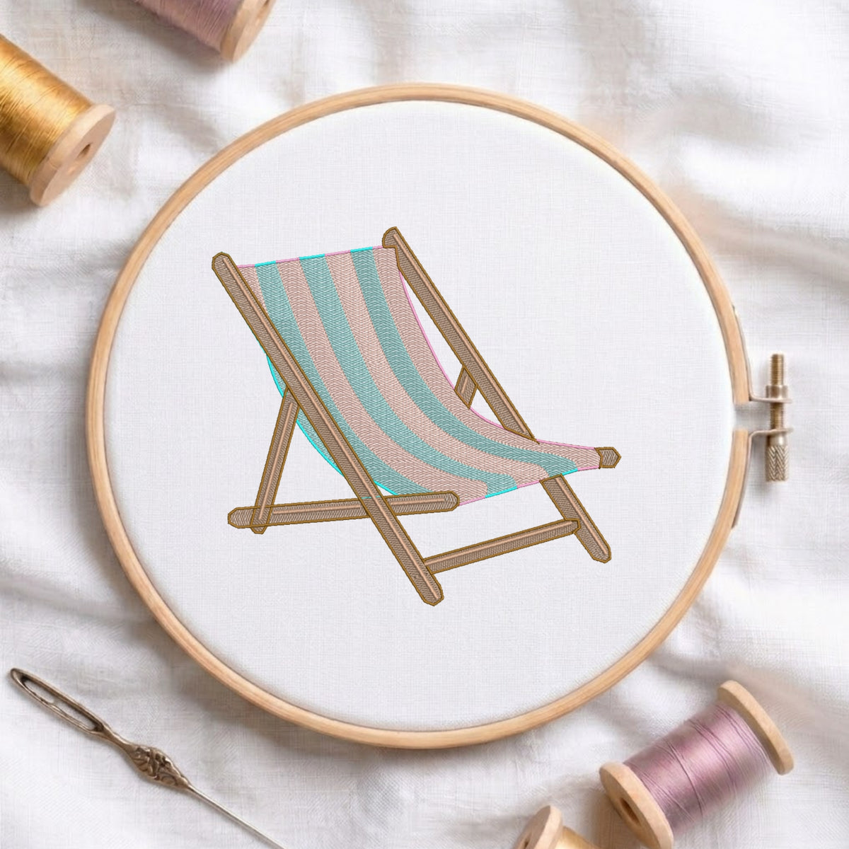 A circular embroidery hoop holding white fabric with a stitched deck chair design in pastel aqua, blush pink, and tan. The hoop is styled on a white fabric background with spools of thread and a needle around it.