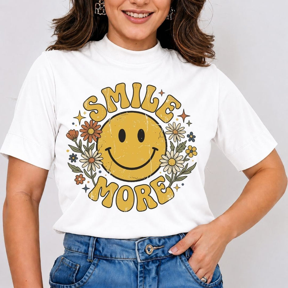 A smiling woman wearing a white mock neck t-shirt printed with a vintage yellow smiley face surrounded by daisies, stars, and the text Smile More.