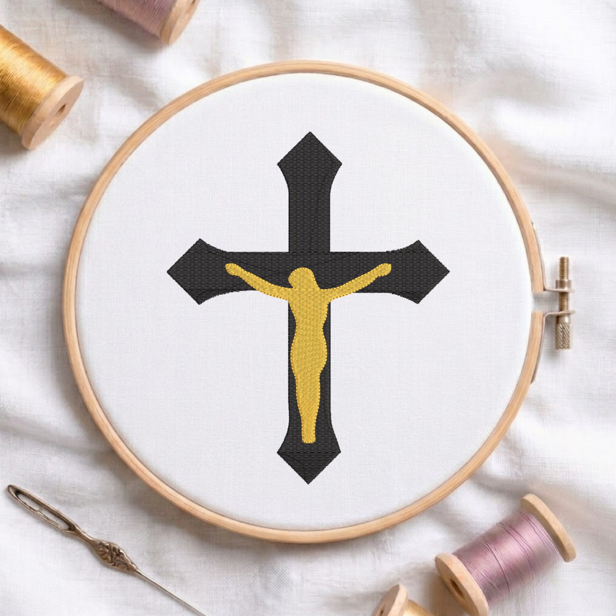A circular embroidery hoop on white fabric displays a black cross with a gold Jesus figure, surrounded by spools of thread and a needle as styled props.