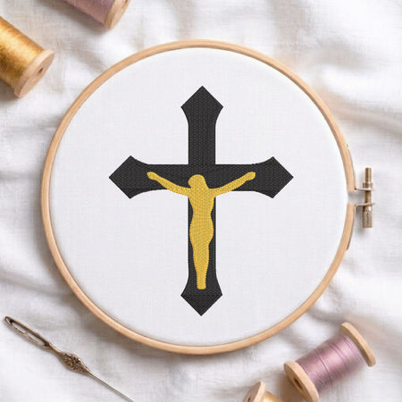 A circular embroidery hoop on white fabric displays a black cross with a gold Jesus figure, surrounded by spools of thread and a needle as styled props.