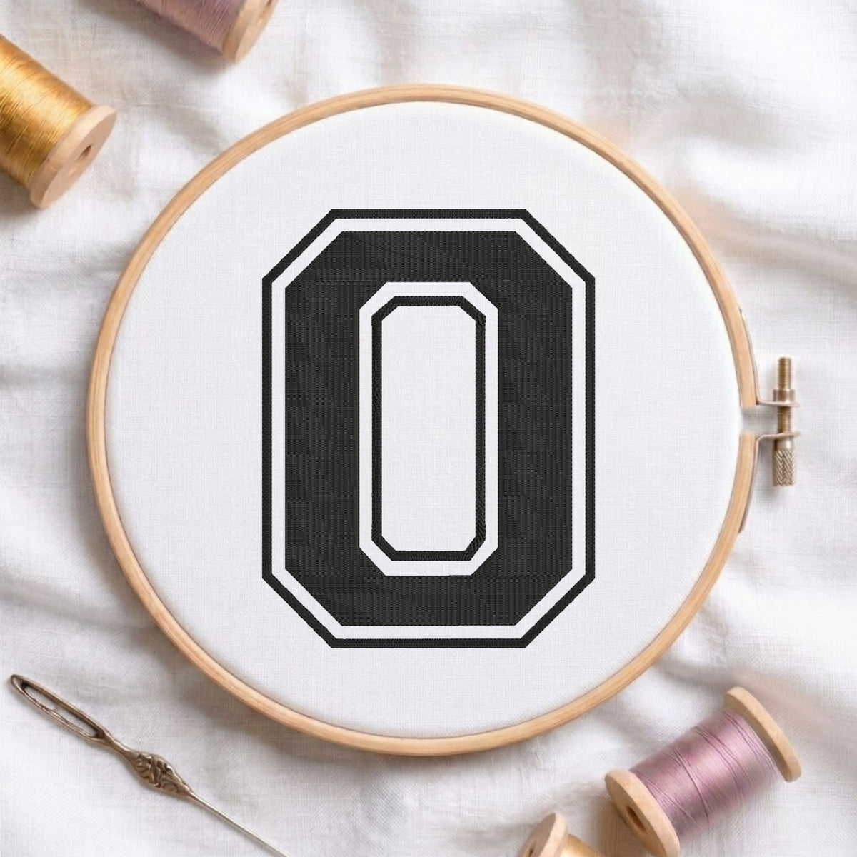 A wooden embroidery hoop holding white fabric with a large black letter O design, styled on a white cloth with spools of thread and a needle.