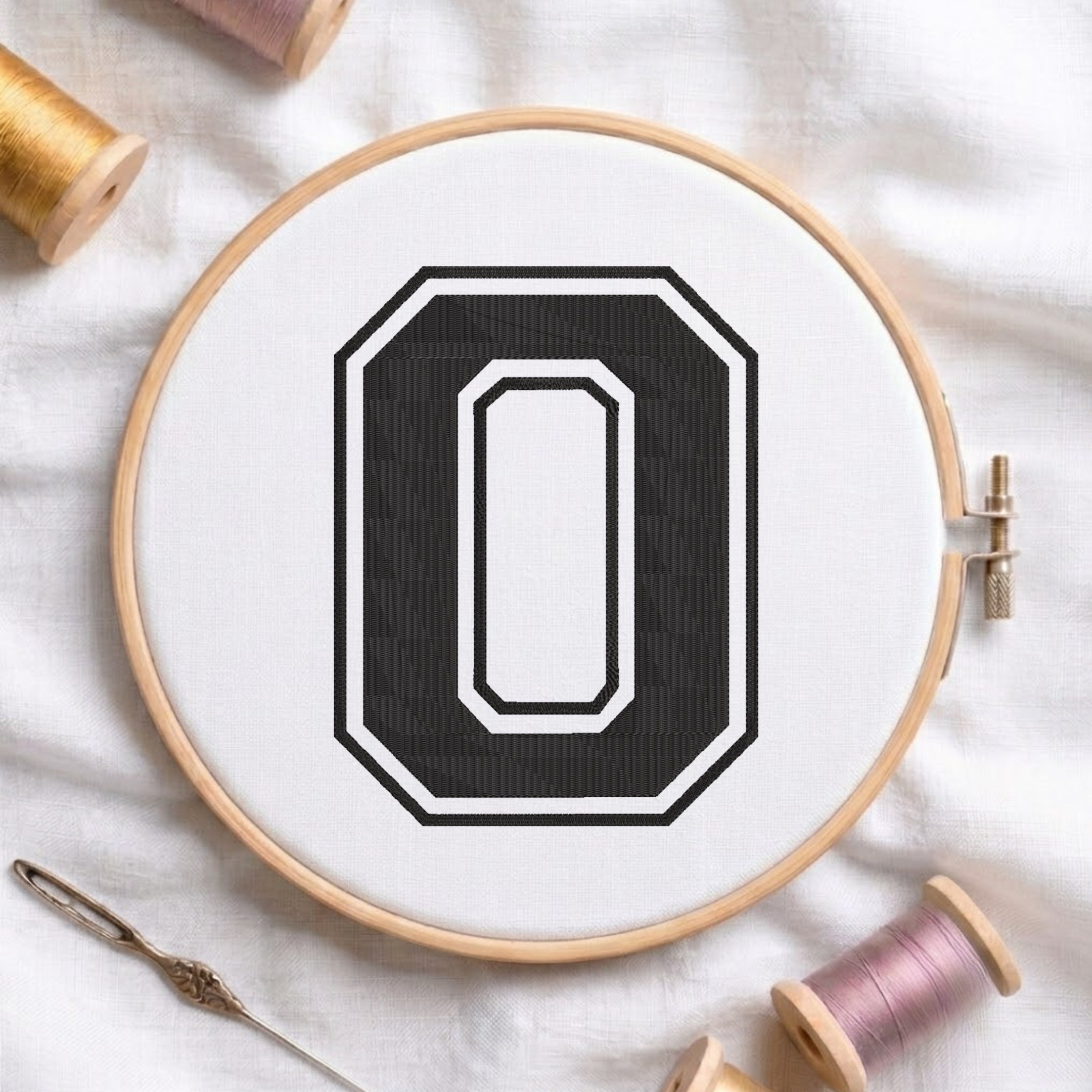 A wooden embroidery hoop holding white fabric with a large black letter O design, styled on a white cloth with spools of thread and a needle.