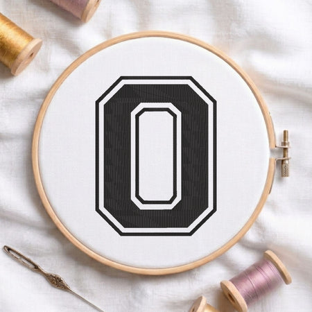 A wooden embroidery hoop holding white fabric with a large black letter O design, styled on a white cloth with spools of thread and a needle.