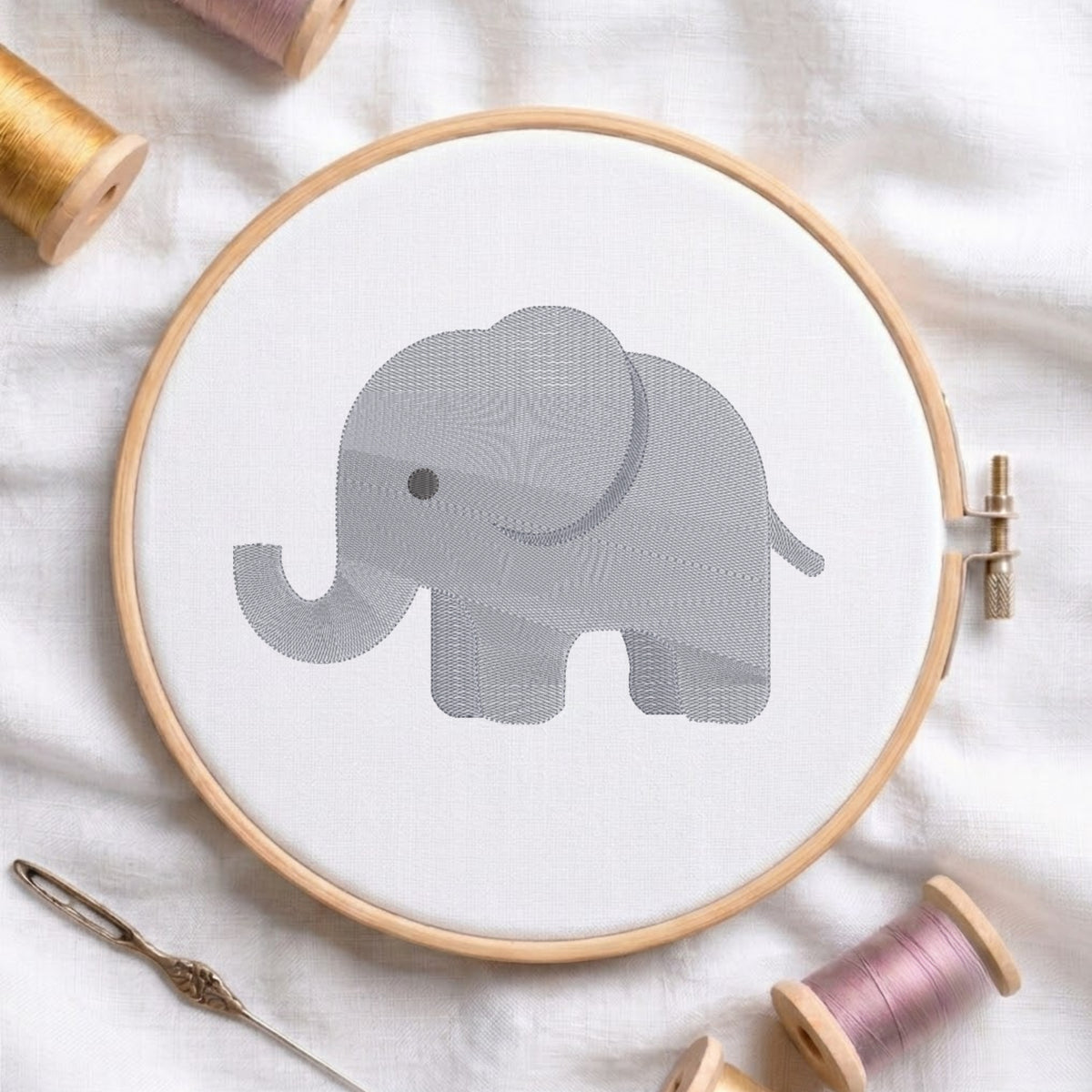 A circular embroidery hoop holding white fabric with a gray elephant design, surrounded by spools of thread and a needle on a white fabric background.