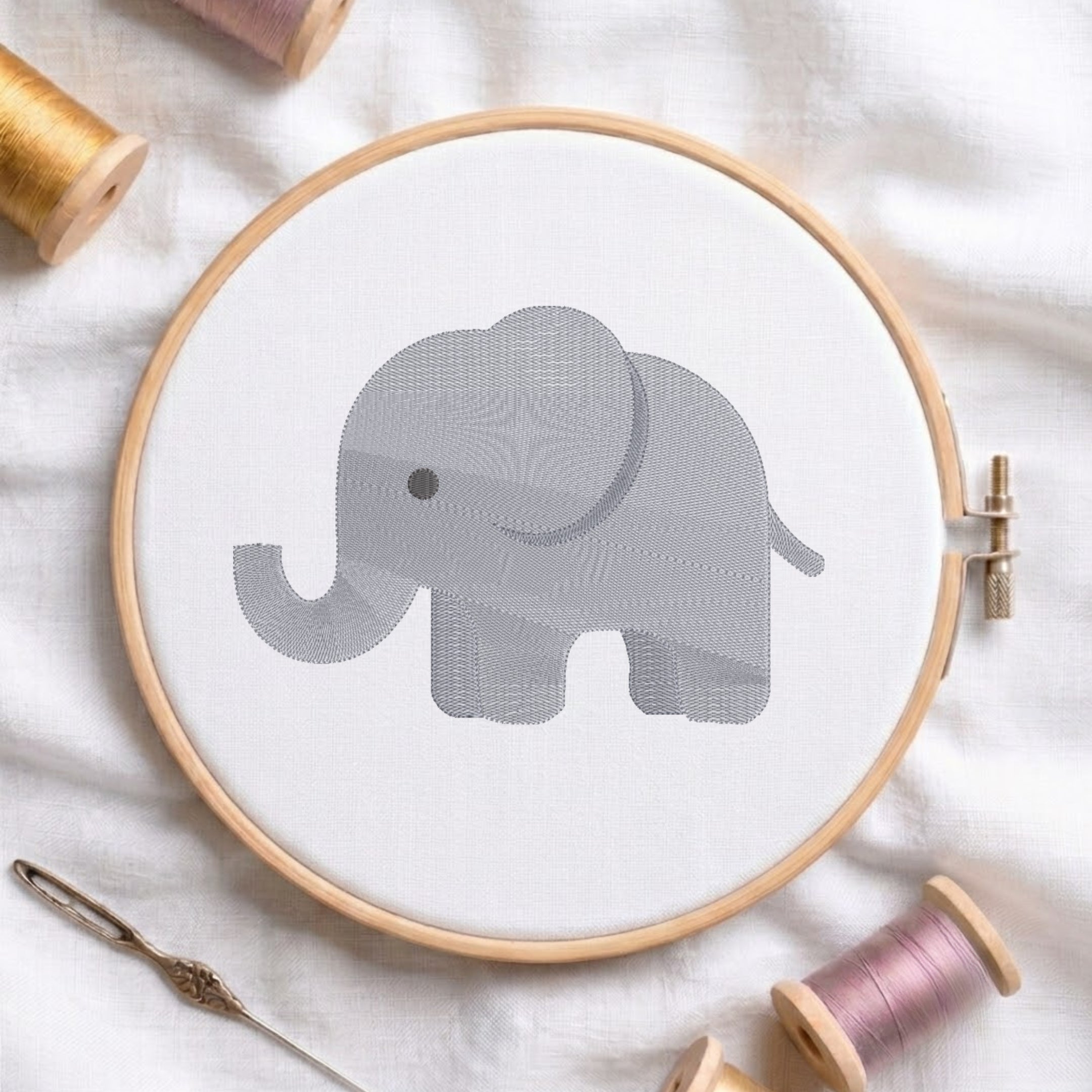A circular embroidery hoop holding white fabric with a gray elephant design, surrounded by spools of thread and a needle on a white fabric background.