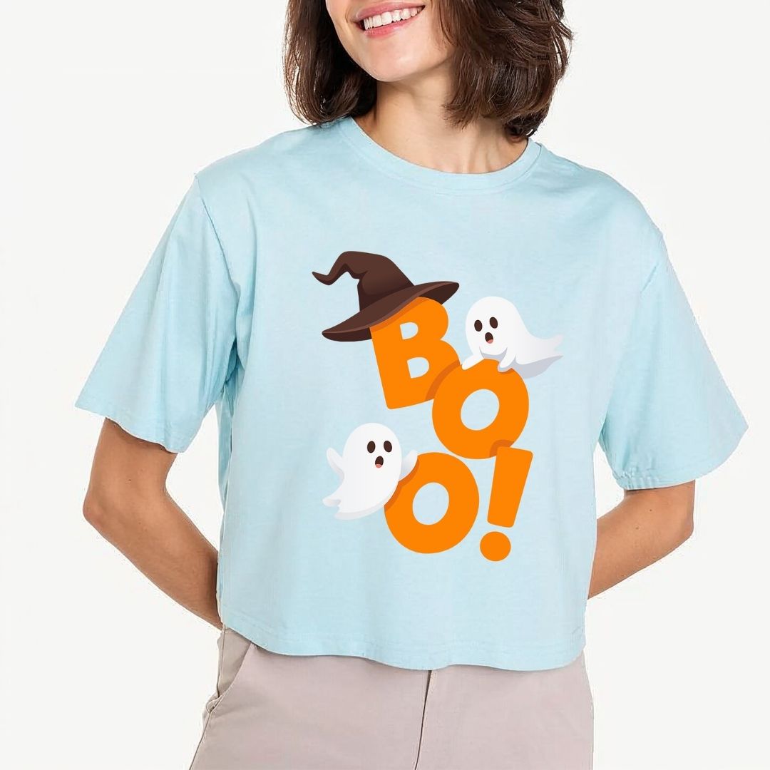 Woman wearing a light blue cropped t-shirt featuring an orange "BOO!" Halloween text design with a witch hat and cute ghosts.