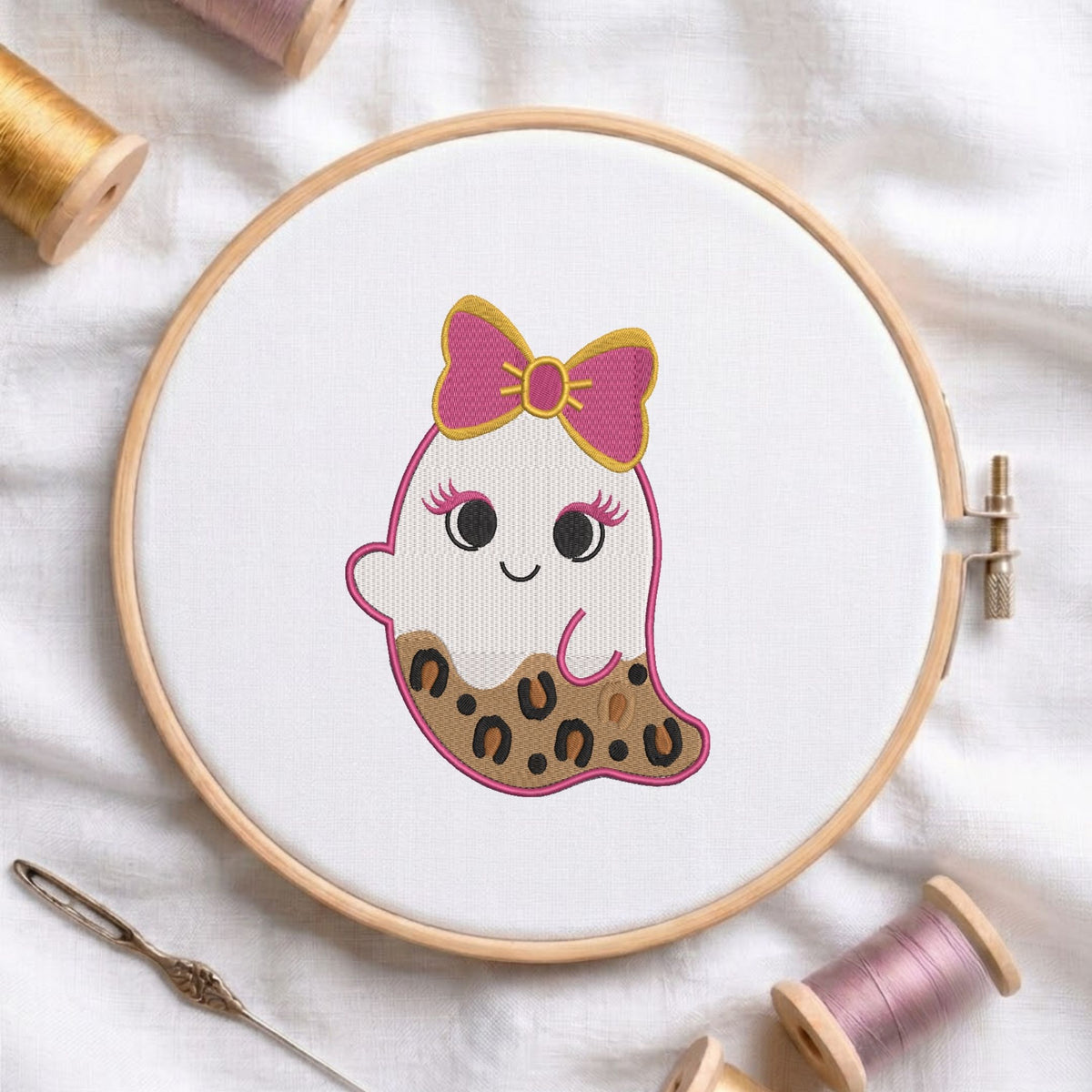 Cute ghost machine embroidery design wearing a pink and gold bow with leopard print details on the lower body, displayed inside a round wooden hoop on a white linen background alongside thread spools and a needle.