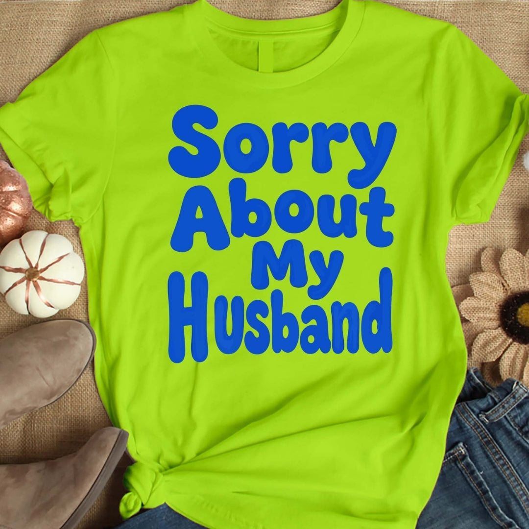 Green t-shirt with blue text 'Sorry About My Husband' on a textured surface.