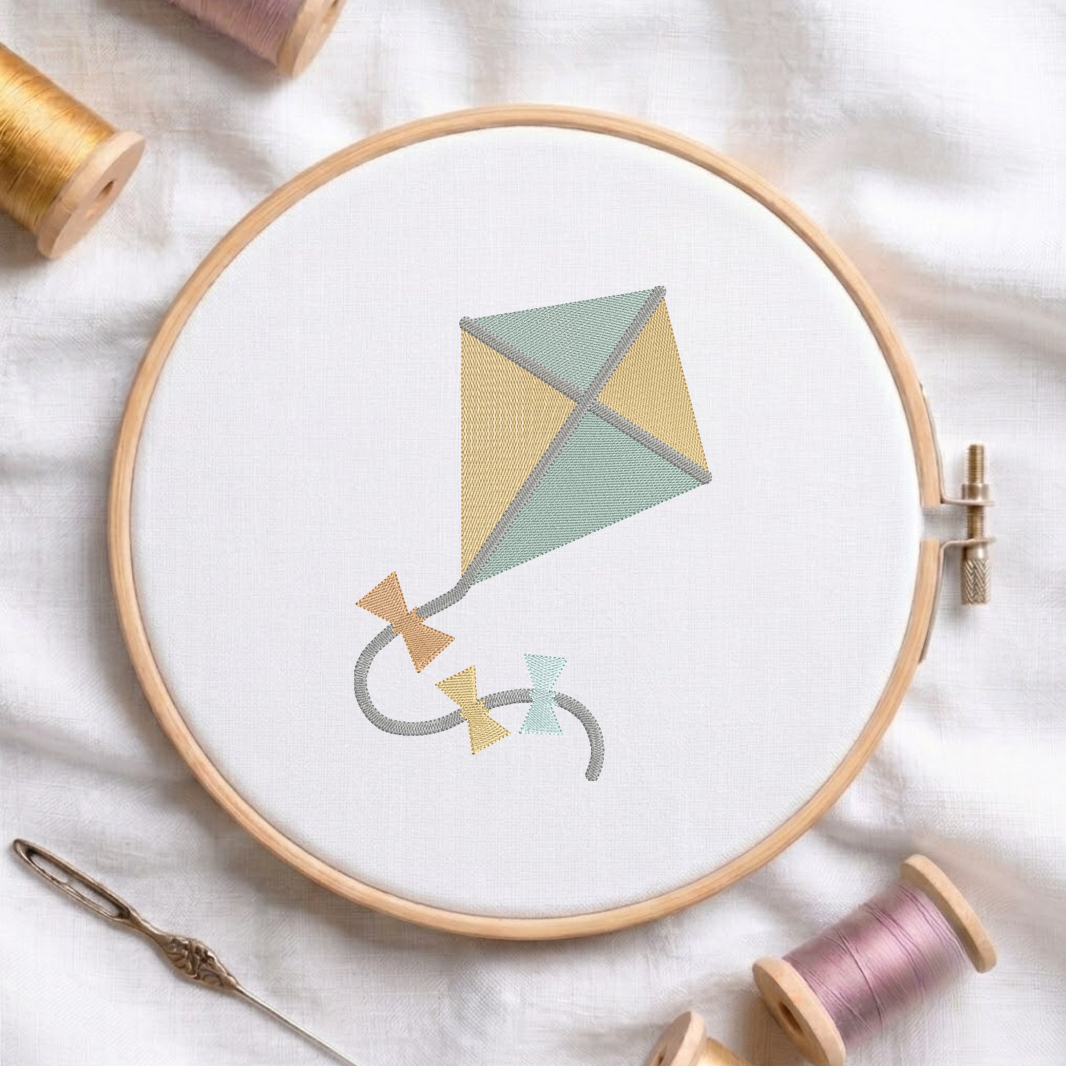 An embroidered cute kite design with yellow and green panels and bow ties on the string, stitched inside a wooden hoop on white fabric with sewing threads.