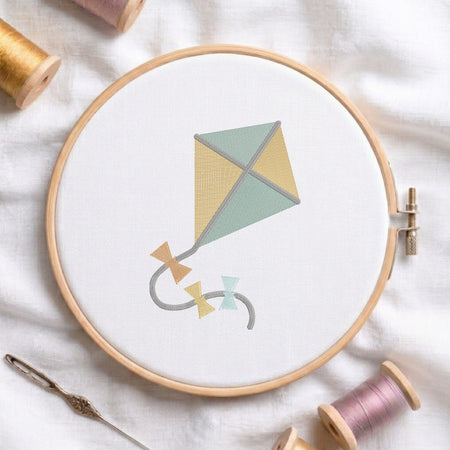 An embroidered cute kite design with yellow and green panels and bow ties on the string, stitched inside a wooden hoop on white fabric with sewing threads.