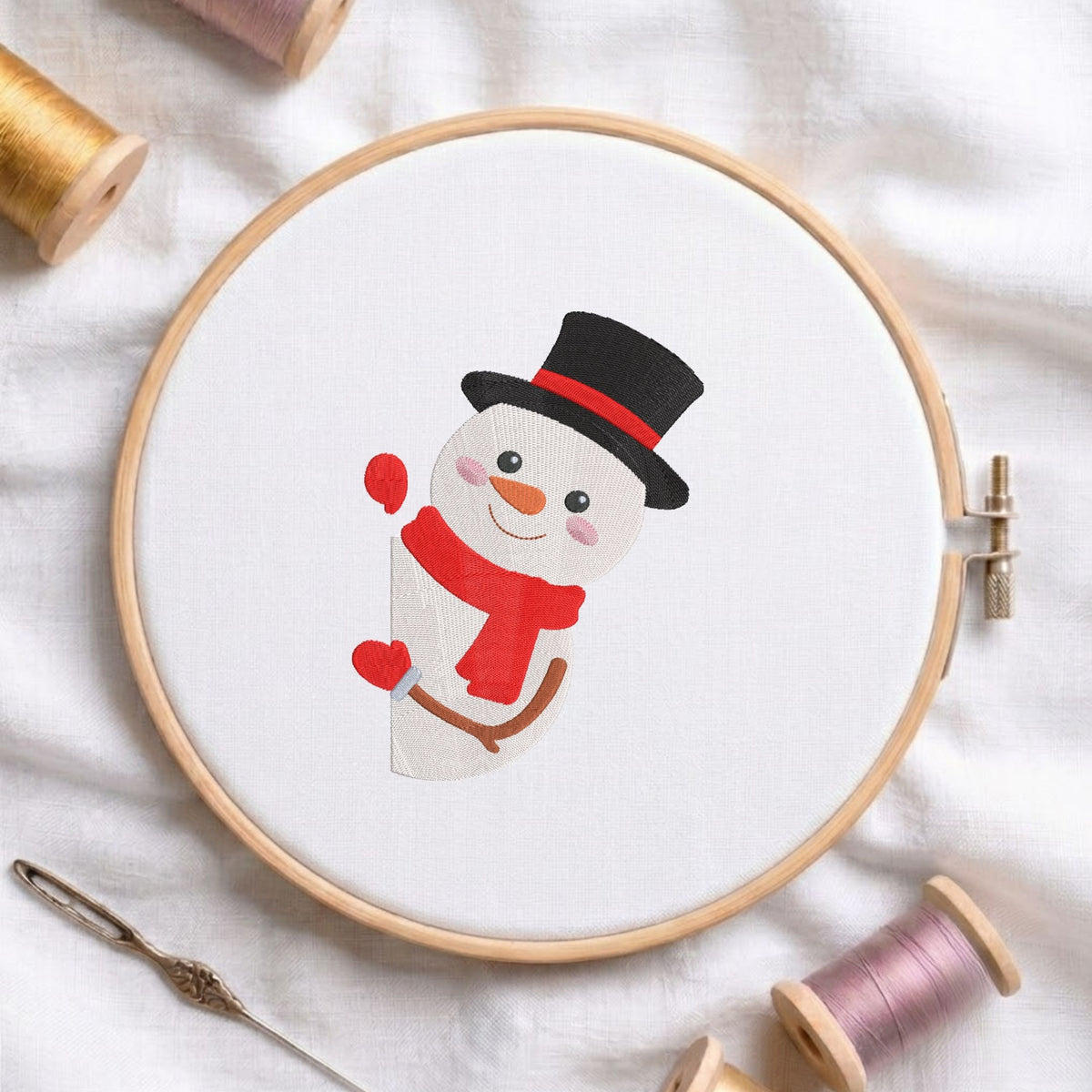 Cute snowman wearing a black top hat, red band, and red scarf machine embroidery design displayed in a wooden hoop on white fabric with thread spools and a needle.