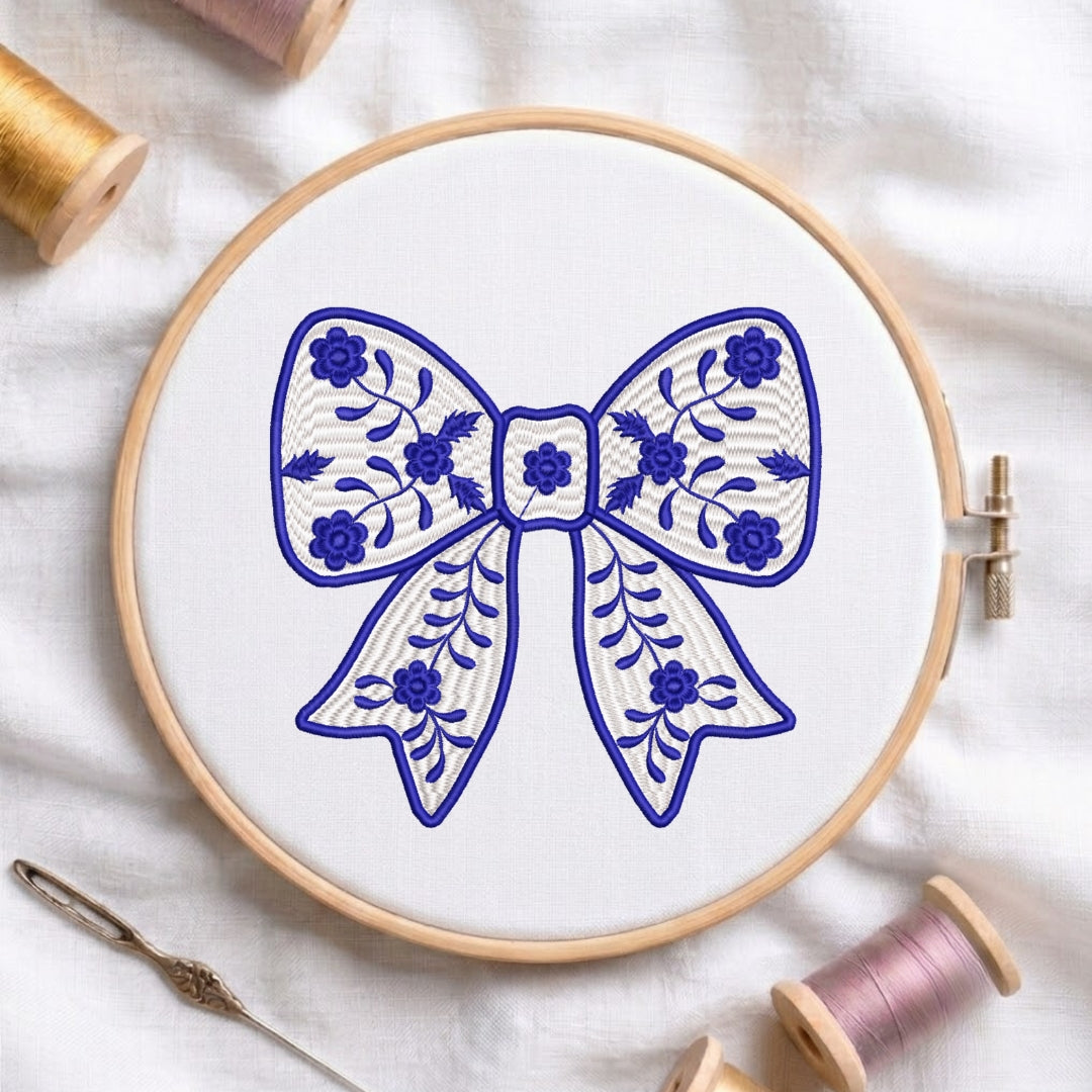 A wooden embroidery hoop holding white fabric with a blue bow design, surrounded by spools of thread and a needle on a white fabric background.