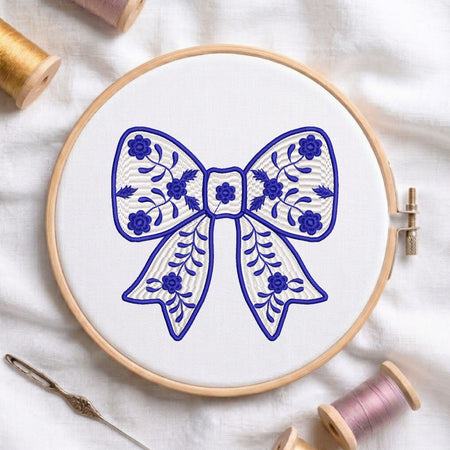A wooden embroidery hoop holding white fabric with a blue bow design, surrounded by spools of thread and a needle on a white fabric background.