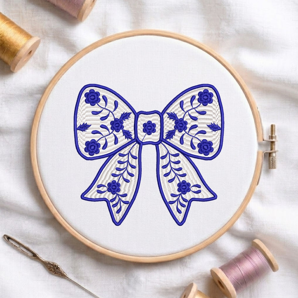 A wooden embroidery hoop holding white fabric with a blue bow design, surrounded by spools of thread and a needle on a white fabric background.