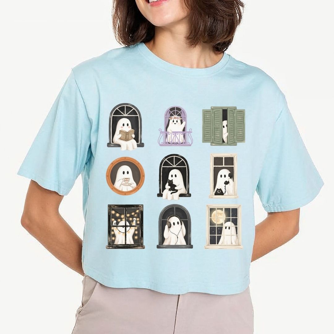 A woman wearing a light blue cropped t-shirt with a graphic print of nine cute cartoon ghosts in various window frames, paired with beige pants against a light gray background.