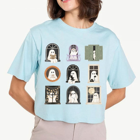 A woman wearing a light blue cropped t-shirt with a graphic print of nine cute cartoon ghosts in various window frames, paired with beige pants against a light gray background.