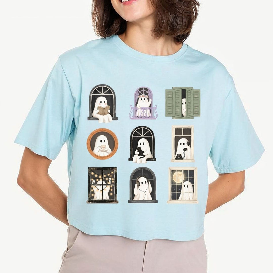 A woman wearing a light blue cropped t-shirt with a graphic print of nine cute cartoon ghosts in various window frames, paired with beige pants against a light gray background.