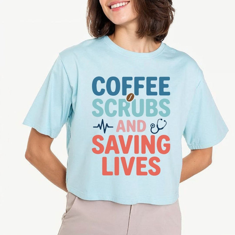 A woman wearing a light blue cropped graphic T-shirt with the text 'COFFEE SCRUBS AND SAVING LIVES' in blue, teal, and coral colors, paired with light beige pants, against a plain off-white background.