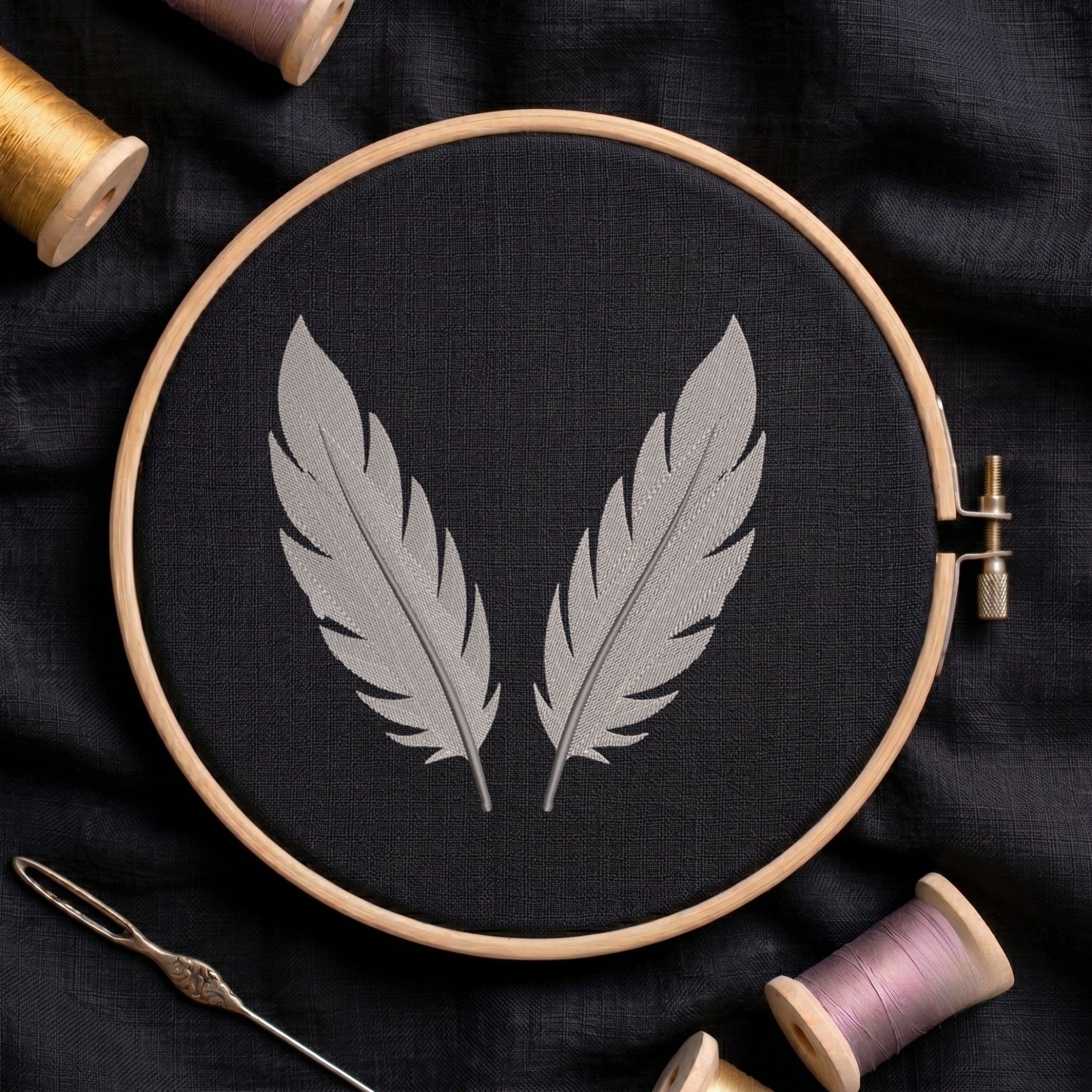 A wooden embroidery hoop holding black fabric with two gray embroidered feathers, surrounded by spools of thread and a needle on a dark fabric background.
