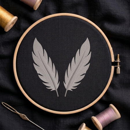 A wooden embroidery hoop holding black fabric with two gray embroidered feathers, surrounded by spools of thread and a needle on a dark fabric background.